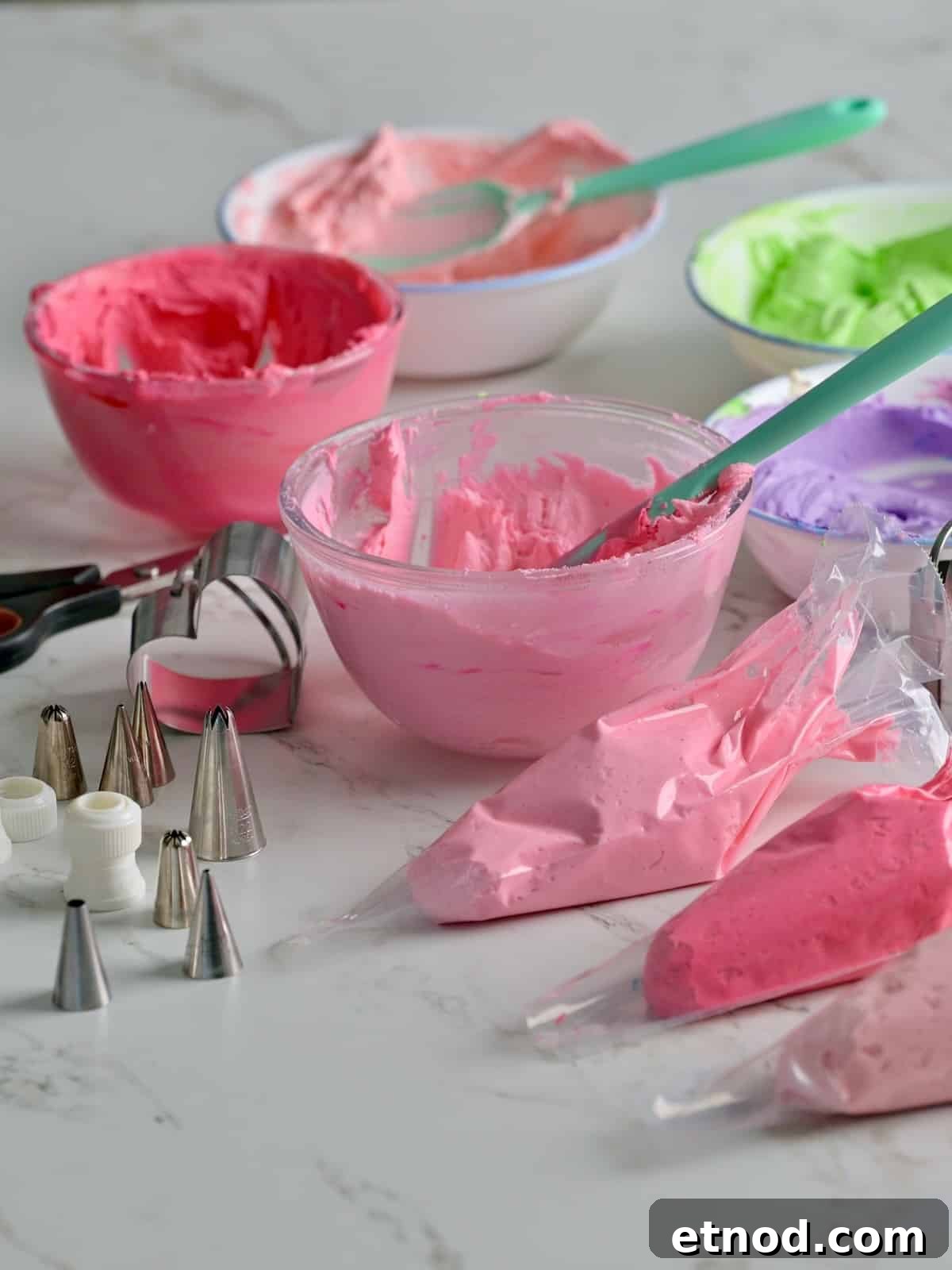 Valentine Sugar Cookies with Creamy Buttercream Frosting 9 3 piping bags filled with light pink, pink, and magenta buttercream, with various bowls of buttercream frosting and piping tips and a heart cookie cutter in view on the table.