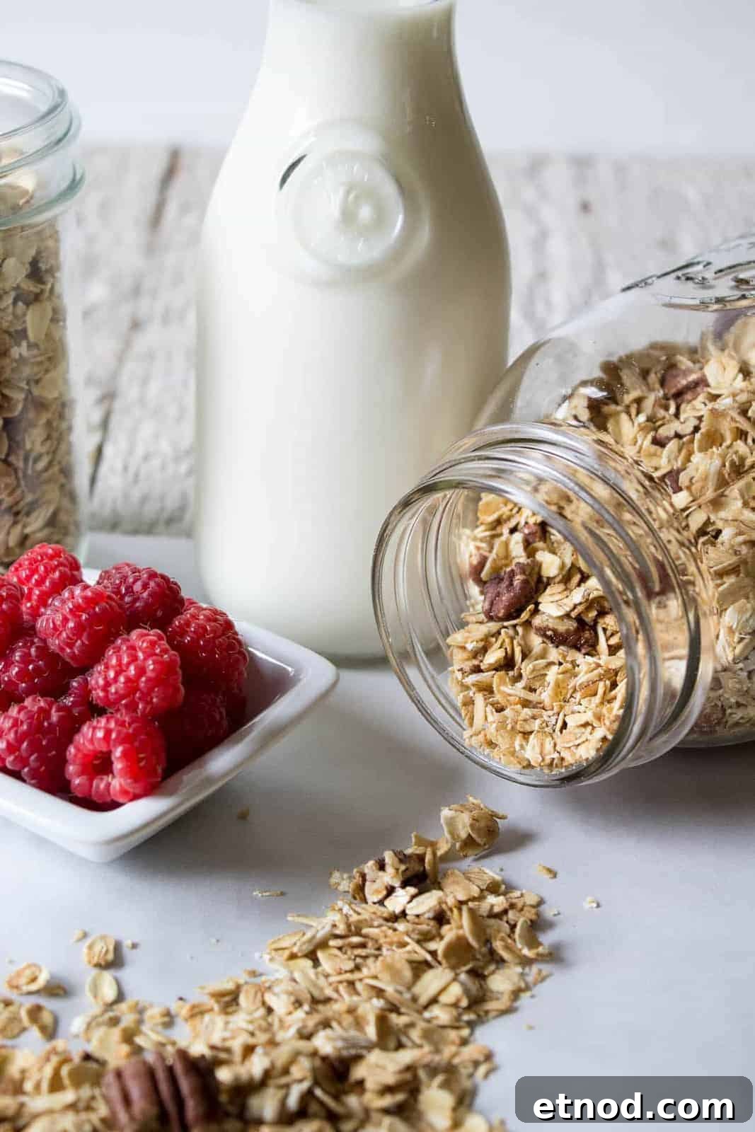 A bottle of milk, a glass jar of rolled oats spilling onto the surface, a jar of oats in the background, and a dish of fresh raspberries—perfect ingredients for make ahead breakfast recipes.