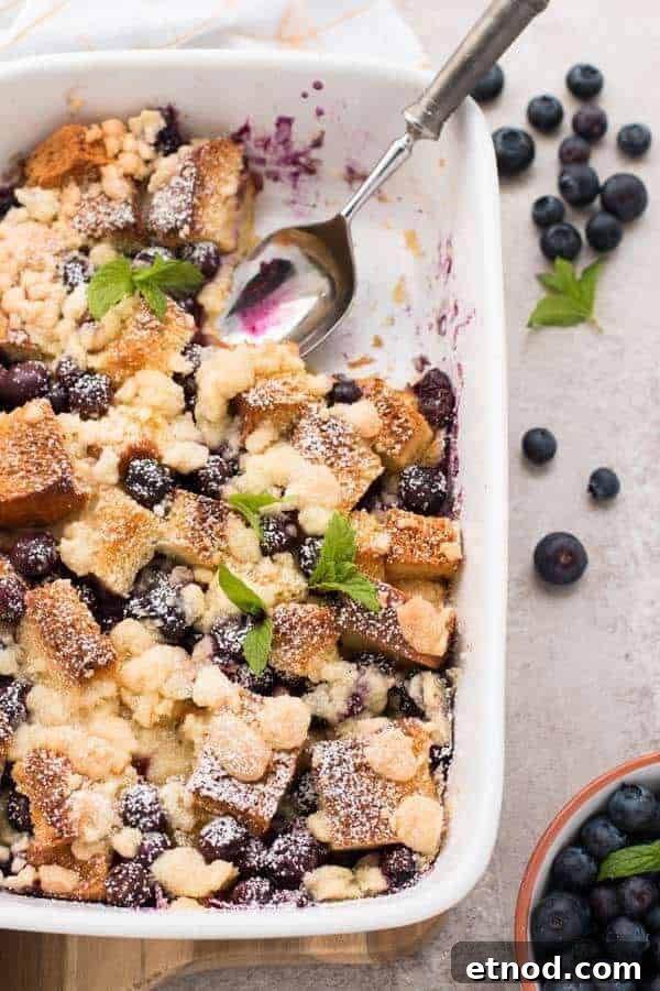 A baked blueberry crumble dessert topped with powdered sugar and mint leaves in a white dish, with a serving spoon and fresh blueberries nearby—perfect for those seeking make ahead breakfast recipes.