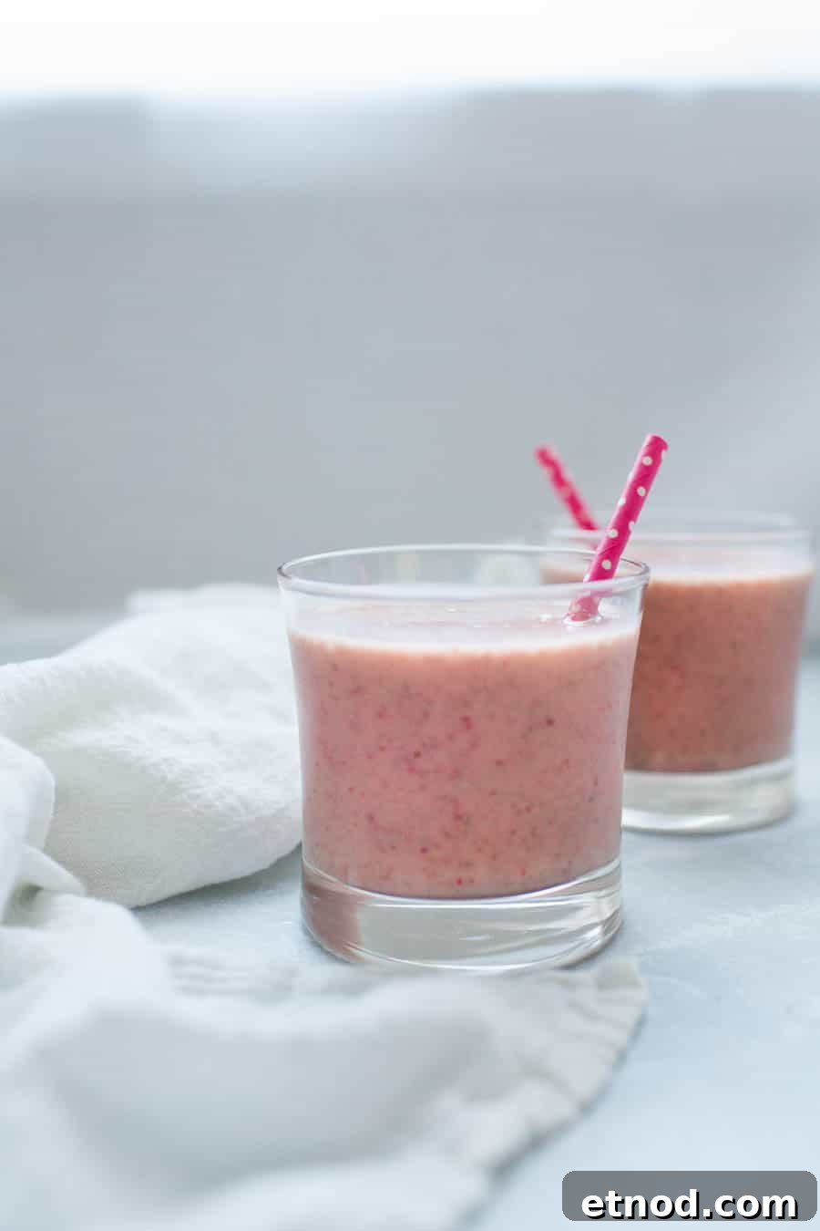 Two glasses of strawberry banana smoothie with straws are placed on a light surface near a white cloth.