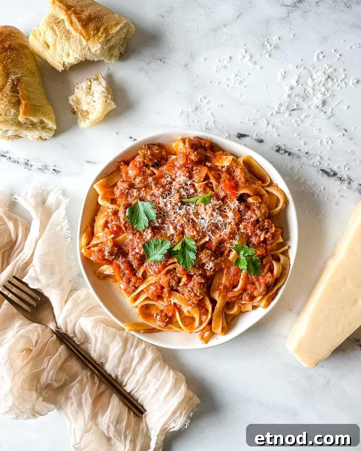 a plate of slow cooker lamb ragu over pappardelle pasta surrounded by a block of parmesan, a white linen, and torn baguette.