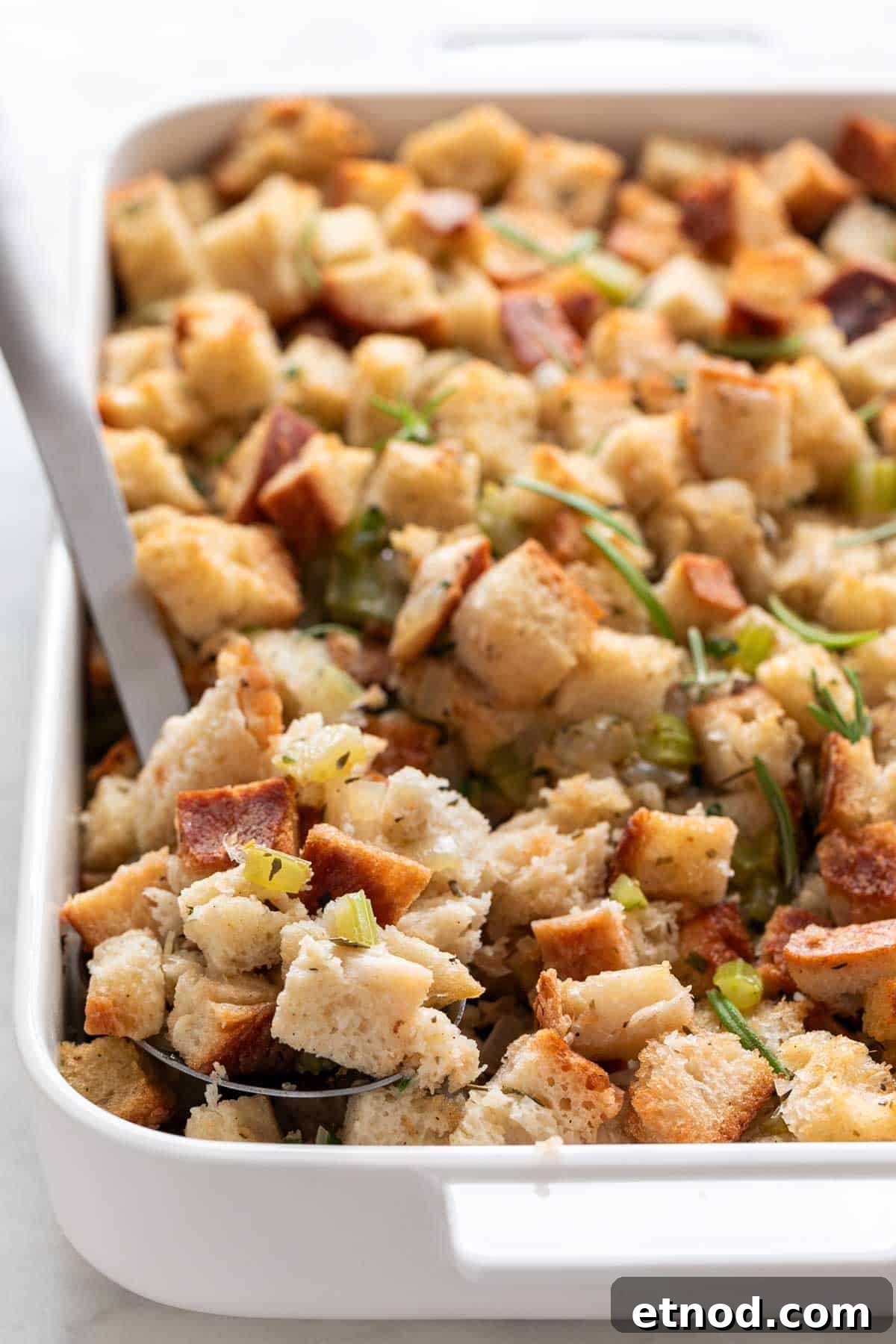 Vegan stuffing in a baking dish with a serving spoon and fresh rosemary on top.