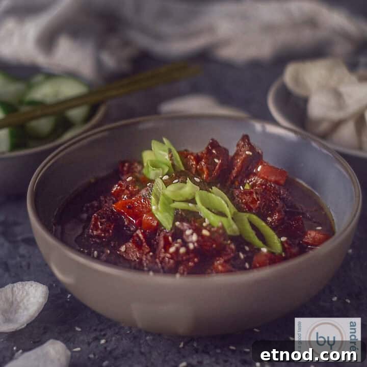 A square picture with a grey bowl filled with slow cooker mongolian beef with spring onion garnish and some prawn crackers.