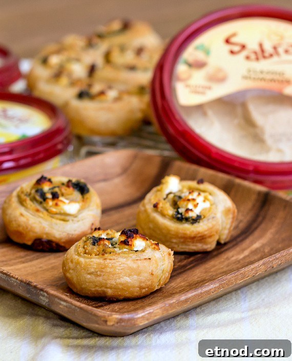 Spinach and Feta Hummus Pinwheels: Party Appetizer 2 3 puff pastry hummus pinwheels on a wooden plate with containers of hummus in the background.