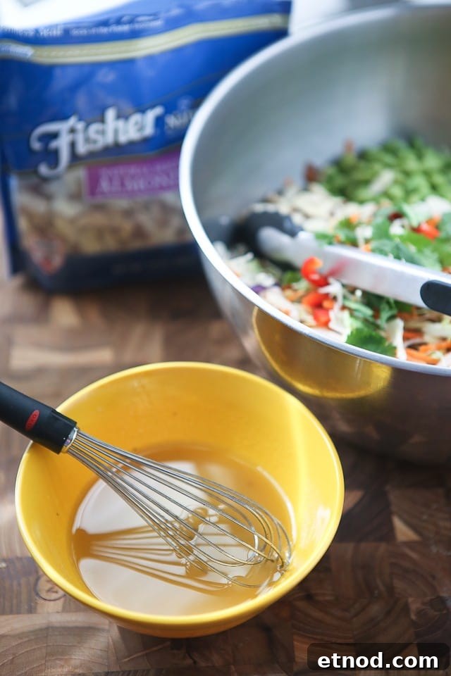 Asian Almond Slaw Salad salad dressing being whisked in a small yellow bowl next to the bowl of salad alongside a package of Fisher almond slices