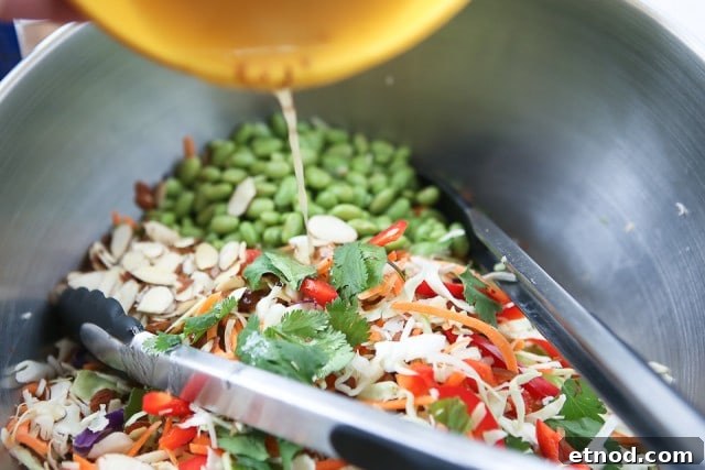 Asian Almond Slaw Salad slaw mix, carrots, red bell peppers, edamamae, almond slices, and cilantro in bowl being covered with dressing