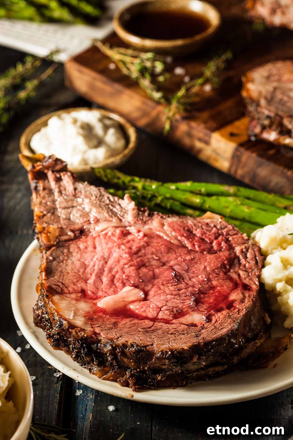 a slice of prime rib on a white plate with mashed potatoes and grilled asparagus, next to a small dish of horseradish sauce. the rest of the standing rib roast is in the background
