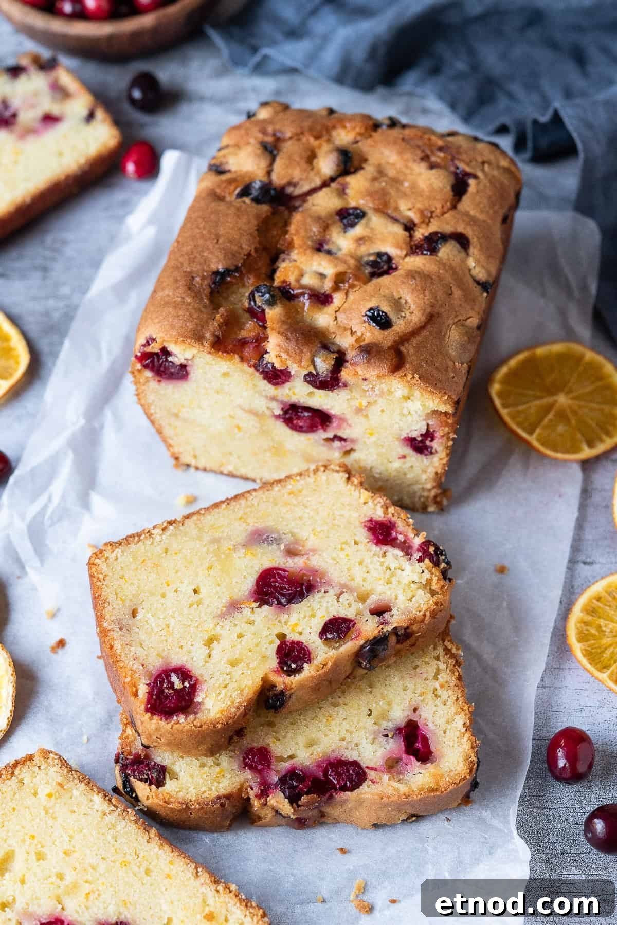 Vegan orange cranberry cake - this delicious loaf cake consists of a vegan orange pound cake studded with tangy fresh cranberries. It is moist, flavourful and perfect for snacking. Eggless and dairy free. Vegan orange cranberry cake on a sheet of white baking parchment with dried orange slices and fresh cranberries.