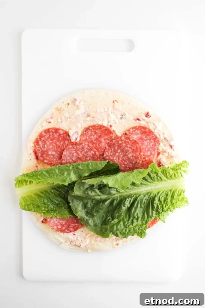 A tortilla wrap on a white cutting board topped with a creamy spread, slices of salami, and romaine lettuce leaves—perfect for making delicious Italian Sub Pinwheels.