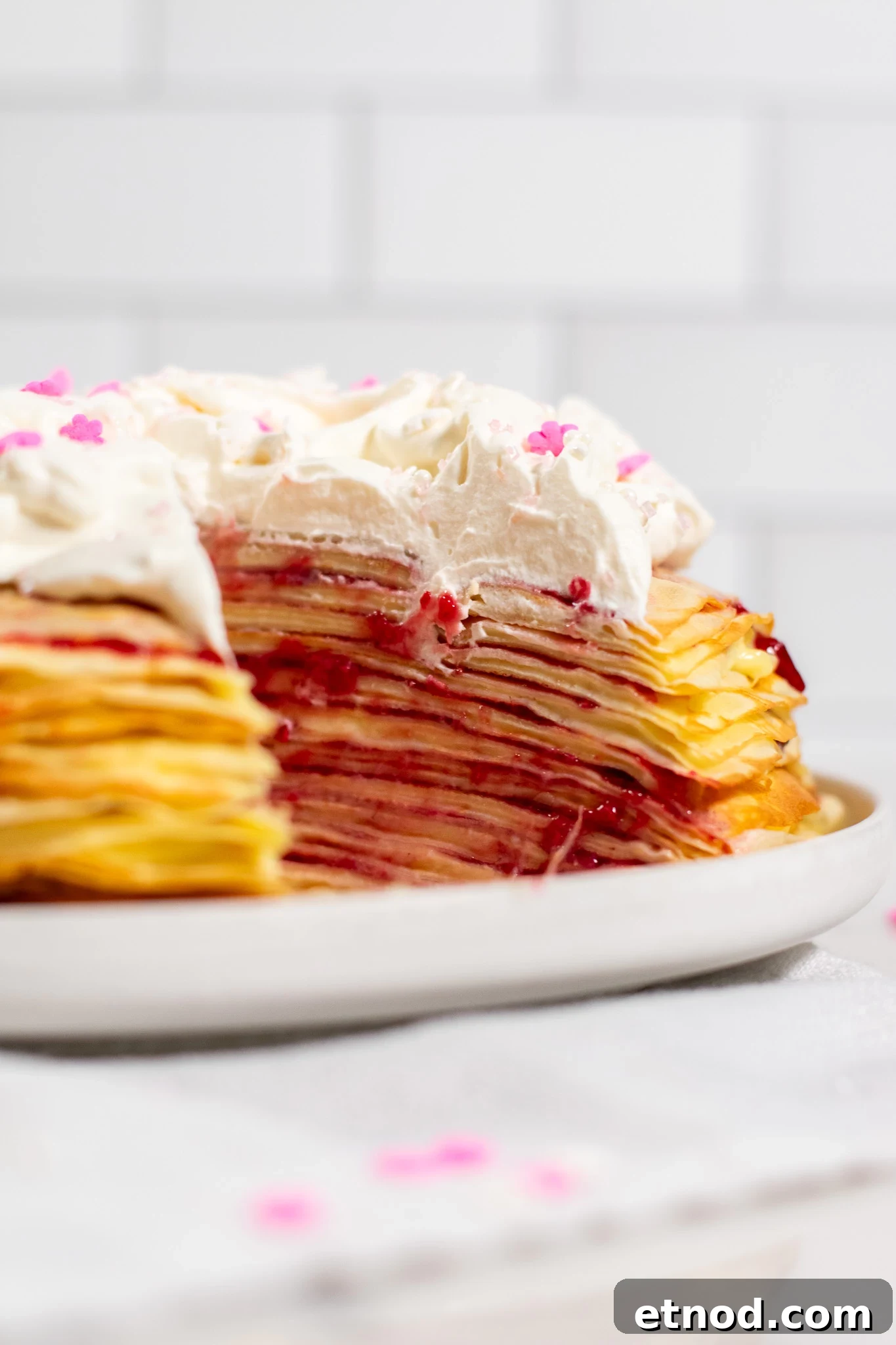 Vanilla and Raspberry Pancake Layer Cake Recipe 6 A pancake layer cake with vanilla pastry cream and raspberry jam, topped with whipped cream, with a slice cut out of it.
