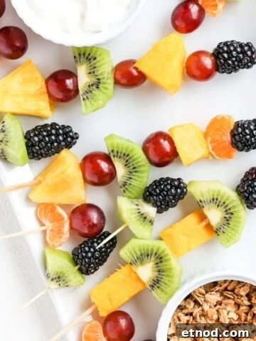Pieces of fresh fruit on toothpicks and bamboo skewers on a white plate with yogurt and granola in separate ramekins.