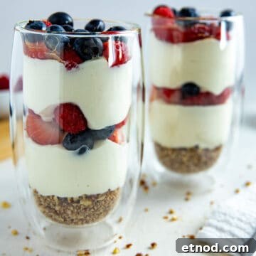 Two tall clear glasses with a granola crust at the bottom, two layers of yogurt and two layers of strawberries and blueberries.