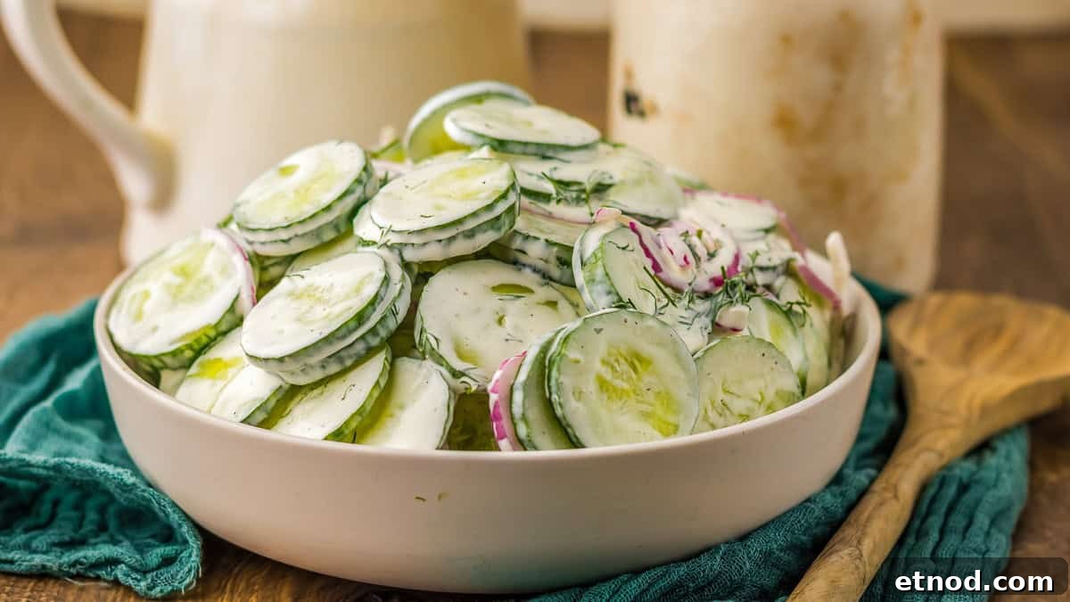 31 Forgotten Grandma Potluck Recipes Worth Bringing Back 17 A bowl of cucumber salad with dill and red onions, dressed in a creamy sauce, on a wooden table beside a wooden spoon and jars.