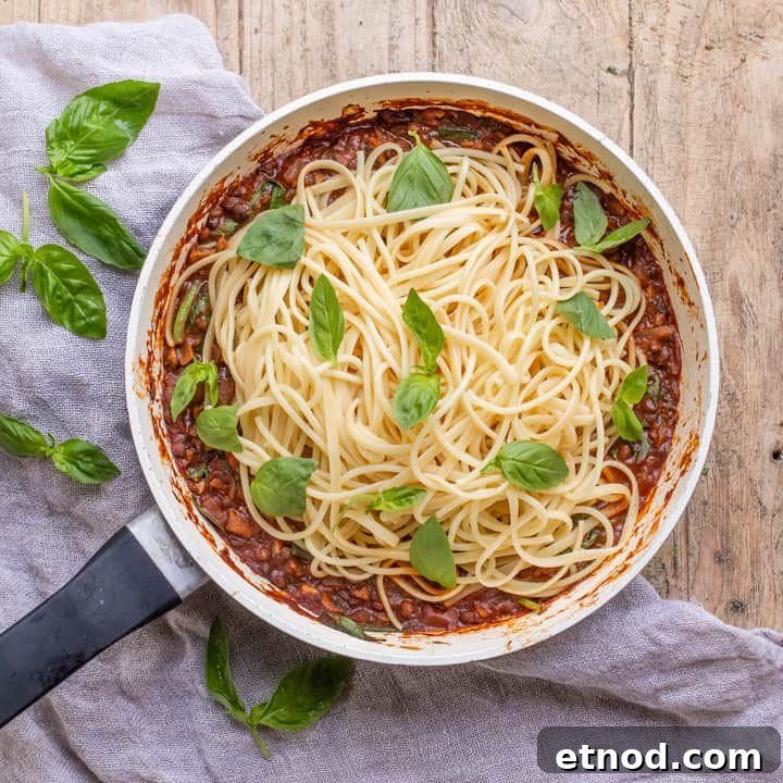 wooden background with white pan with vegan spaghetti bolognese piled up and scattered with fresh basil leaves