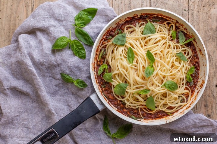 wooden background with white pan with vegan spaghetti bolognese piled up and scattered with fresh basil leaves