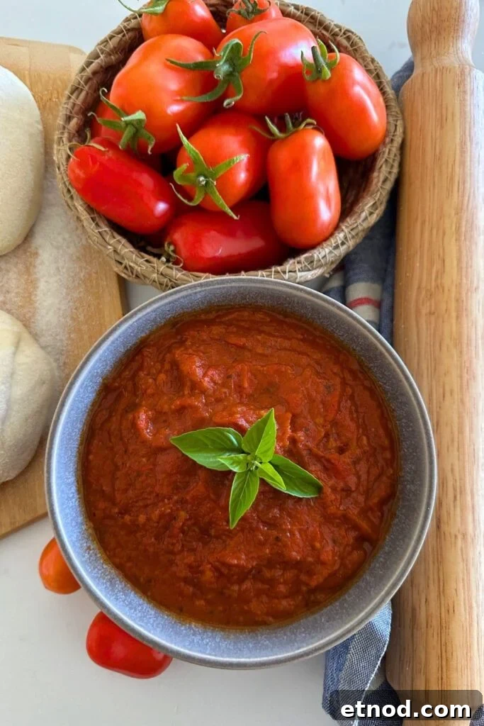 Authentic Homemade Pizza Sauce Recipe for Restaurant-Style Flavor 3 Homemade pizza sauce made using fresh roma tomatoes. The pizza sauce is displayed in a grey stoneware bowl with a piece of green basil in the centre. You can also see some pizza dough, a bowl of roma tomatoes and a rolling pin in the photo.