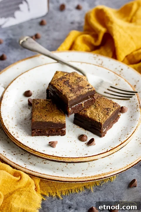4 salted caramel brownies in a stack on top of 3 stacked plates with a fork. Set on a yellow linen napkin over a grey background, there's also scattered chocolate chips and a Bad Brownie box in the background.