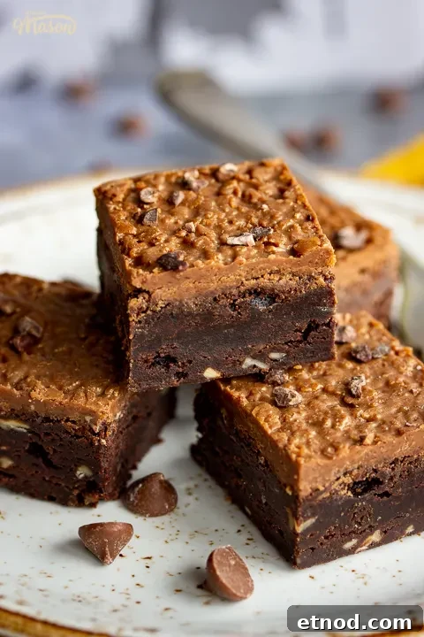 Close up of 4 Ferrero Rocher brownies and a fork on top of 3 stacked plates. Set against a grey backdrop, there's also a Bad Brownie box, a yellow linen napkin and chocolate chips in the background.
