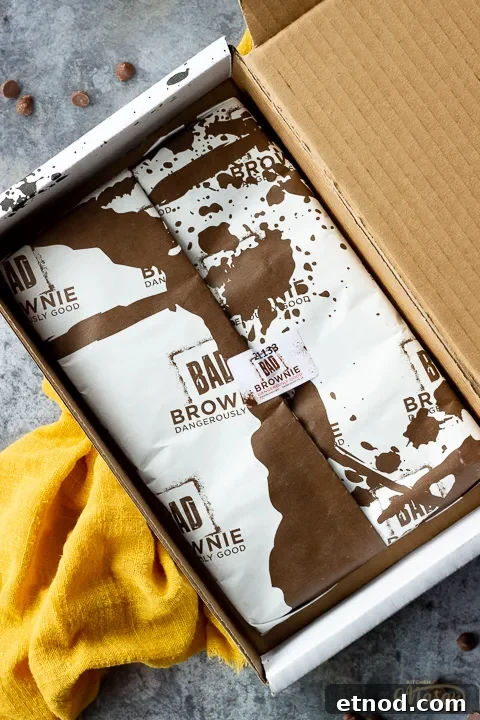 An open but still wrapped box of Bad Brownie brownies on a yellow linen napkin against a grey backdrop with chocolate chips scattered around.