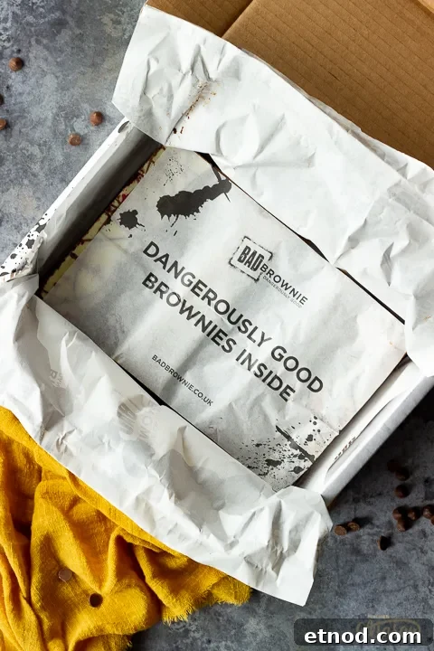 An open box of Bad Brownie brownies with a sheet saying "dangerously good brownies inside" on a yellow linen napkin against a grey backdrop with chocolate chips scattered around.