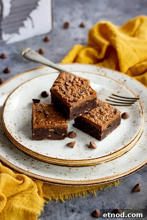 4 Ferrero Rocher brownies in a stack on top of 3 stacked plates with a fork. Set on a yellow linen napkin over a grey background, there's also scattered chocolate chips and a Bad Brownie box in the background.