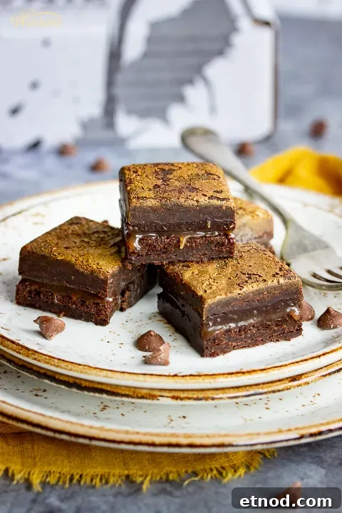 Close up of 4 salted caramel brownies and a fork on top of 3 stacked plates. Set against a grey backdrop, there's also a Bad Brownie box, a yellow linen napkin and chocolate chips in the background.