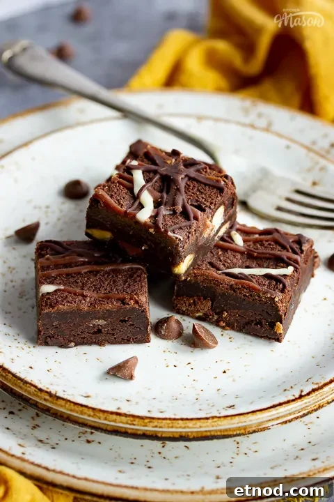 Close up of 4 triple chocolate brownies and a fork on top of 3 stacked plates. Set against a grey backdrop, there's also a Bad Brownie box, a yellow linen napkin and chocolate chips in the background.