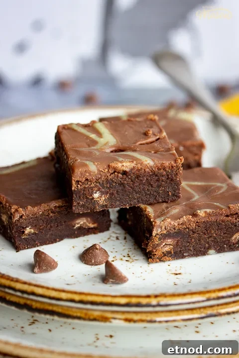 Close up of 4 chocolate orange brownies and a fork on top of 3 stacked plates. Set against a grey backdrop, there's also a Bad Brownie box, a yellow linen napkin and chocolate chips in the background.