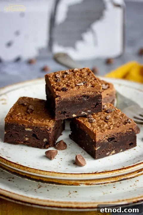 Close up of 4 Ferrero Rocher brownies and a fork on top of 3 stacked plates. Set against a grey backdrop, there's also a Bad Brownie box, a yellow linen napkin and chocolate chips in the background.