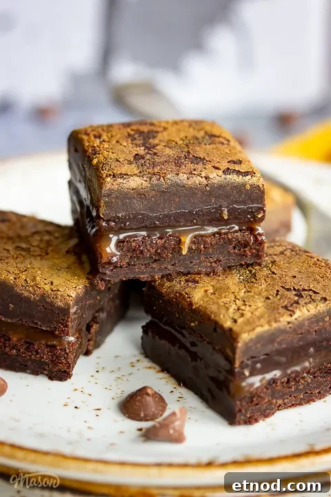 Close up of 4 salted caramel brownies and a fork on top of 3 stacked plates. Set against a grey backdrop, there's also a Bad Brownie box, a yellow linen napkin and chocolate chips in the background.