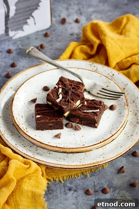 4 triple chocolate brownies in a stack on top of 3 stacked plates with a fork. Set on a yellow linen napkin over a grey background, there's also scattered chocolate chips and a Bad Brownie box in the background.