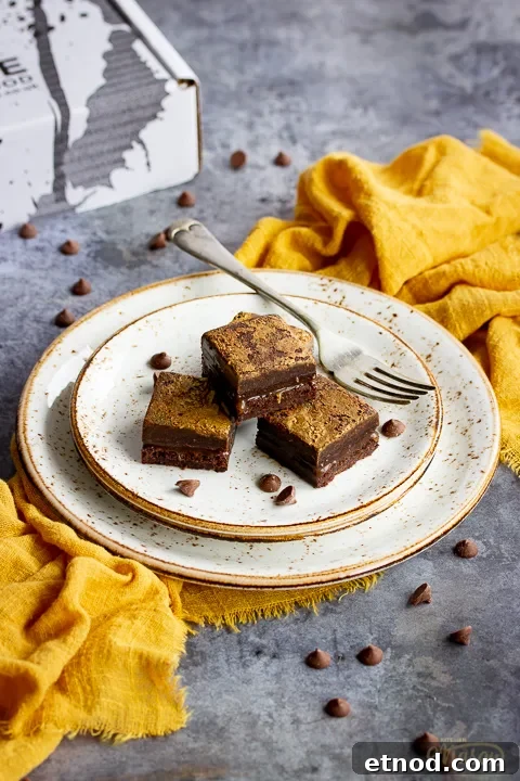 4 salted caramel brownies in a stack on top of 3 stacked plates with a fork. Set on a yellow linen napkin over a grey background, there's also scattered chocolate chips and a Bad Brownie box in the background.