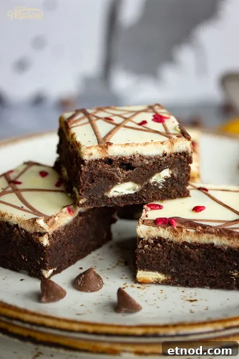 Close up of 4 white chocolate raspberry brownies and a fork on top of 3 stacked plates. Set against a grey backdrop, there's also a Bad Brownie box, a yellow linen napkin and chocolate chips in the background.
