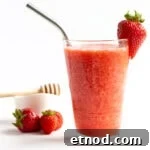 Glass of strawberry slushie with small bowl of honey and a few strawberries in the background.