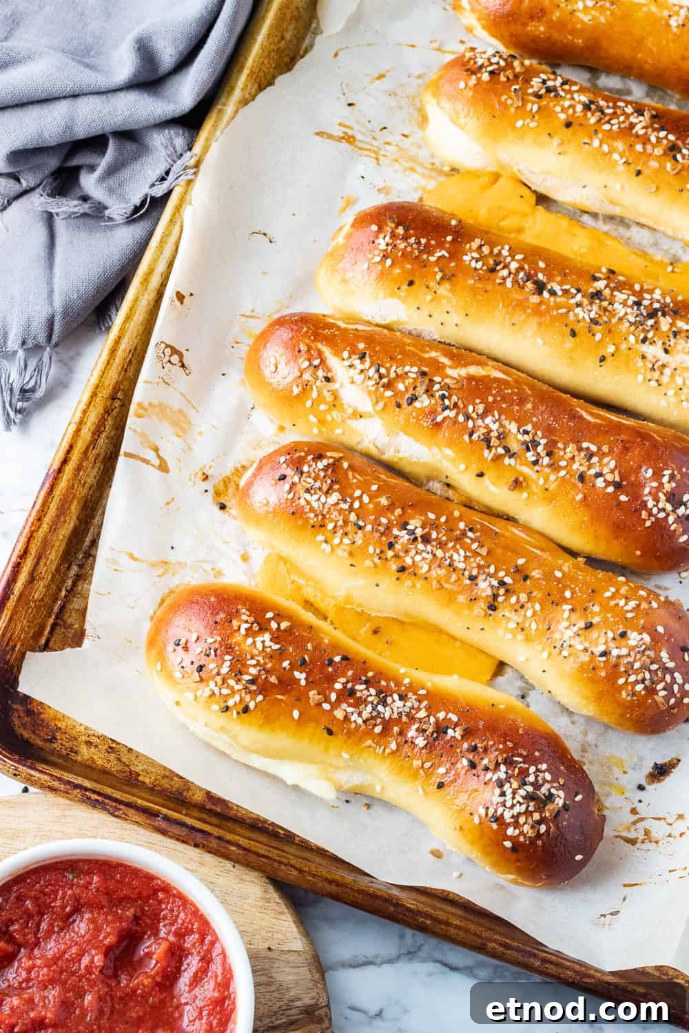 Easy cheese stuffed pretzel with homemade soft pretzel dough, stuffed with cheddar cheese and crunchy seasonings on top. Baked cheese stuffed pretzels on pan with towel and bowl of pizza sauce.