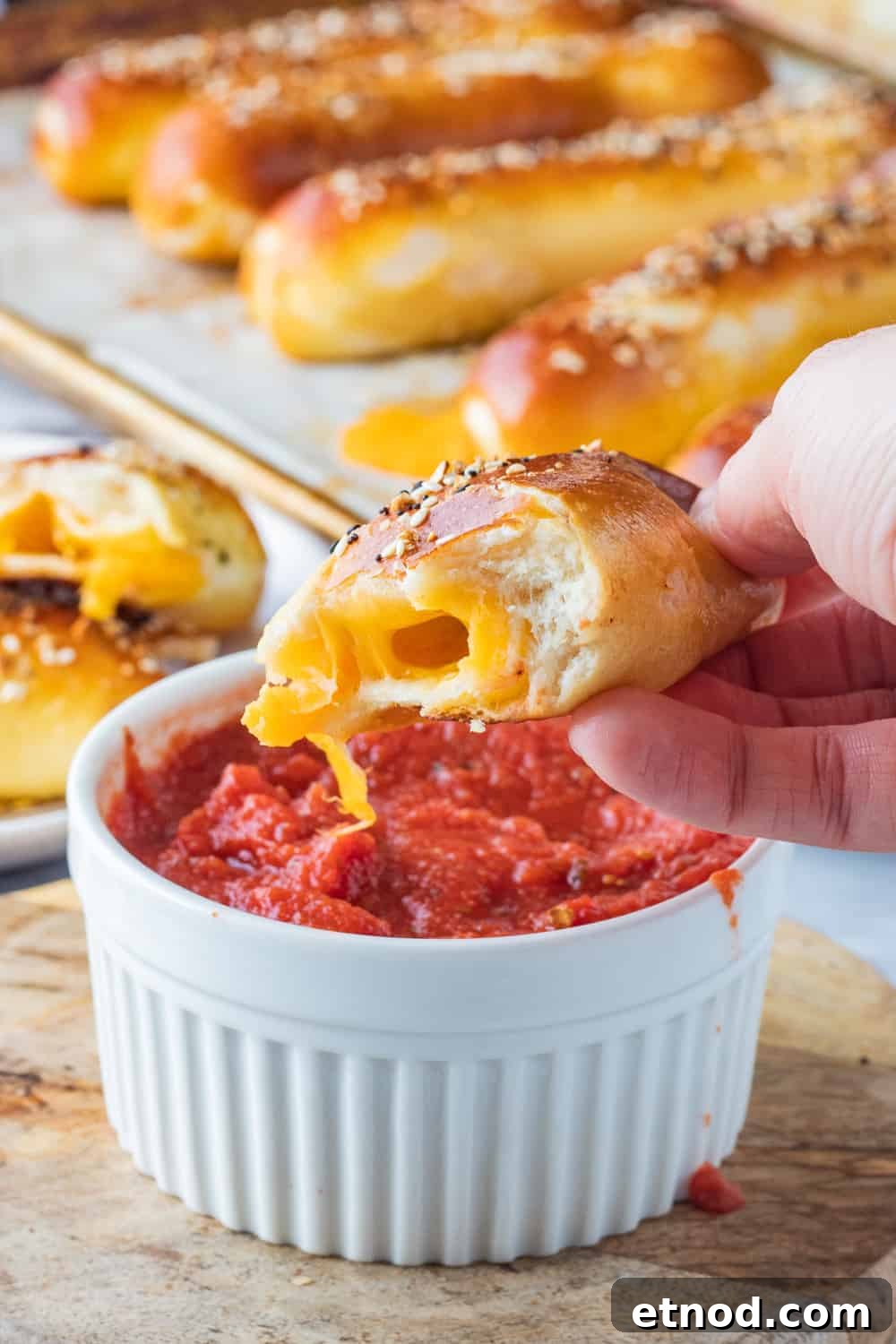 Easy cheese stuffed pretzel with homemade soft pretzel dough, stuffed with cheddar cheese and crunchy seasonings on top. Half of a baked cheese pretzel above white bowl of pizza sauce.