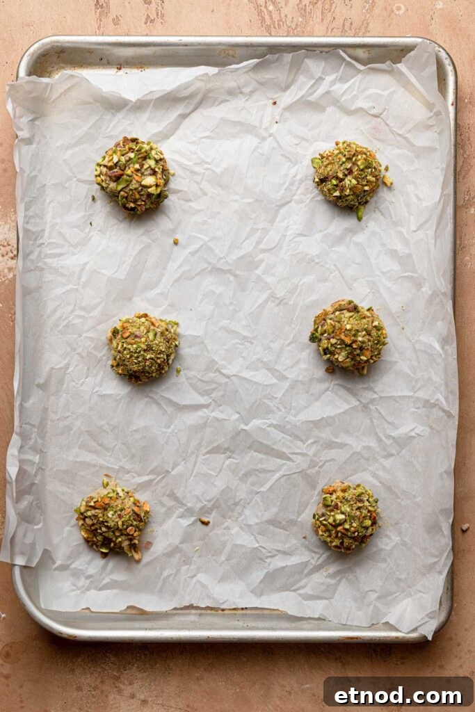 A pan lined with parchment paper and topped with six pistachio chocolate chipe dough balls.