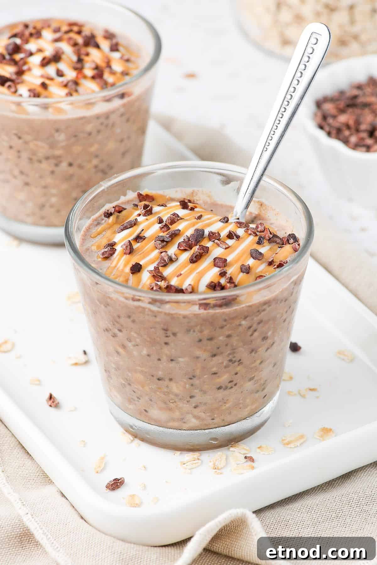 Jar of overnight oats, sitting on a white plate, garnished with peanut butter and cacao nibs, with a spoon in the jar.