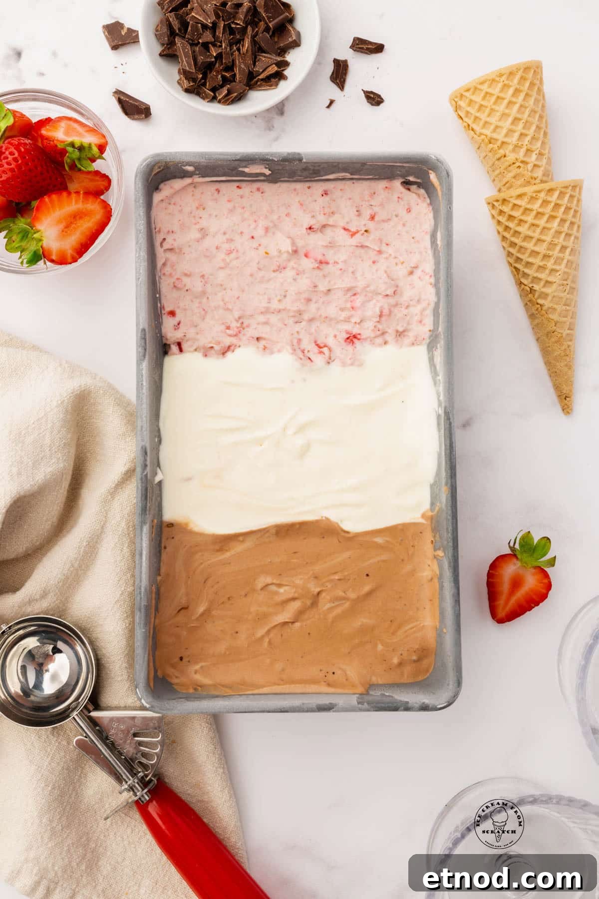 Homemade Neapolitan Ice Cream: Vanilla, Chocolate and Strawberry 5 Strawberry, vanilla, and chocolate ice cream, layered into a metal loaf pan, viewed from above. Next to the pan of ice cream are two sugar cones, a bowl of strawberries, a bowl of chocolate chunks, and a red and silver ice cream scoop.