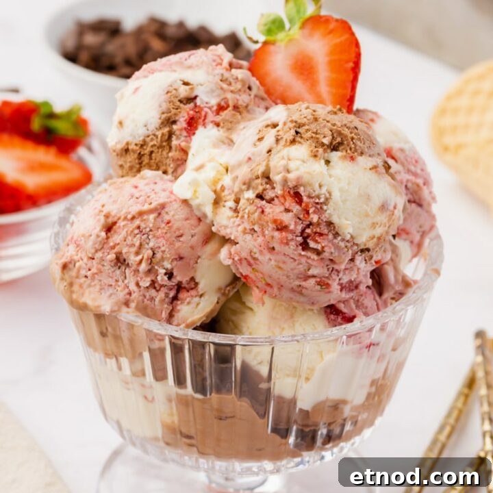 homemade no churn neapolitan ice cream, scooped into a glass, ridged, footed ice cream dish. Two spoons are on the table next to the glass. Dishes of sliced strawberries and chocolate chips are in the background
