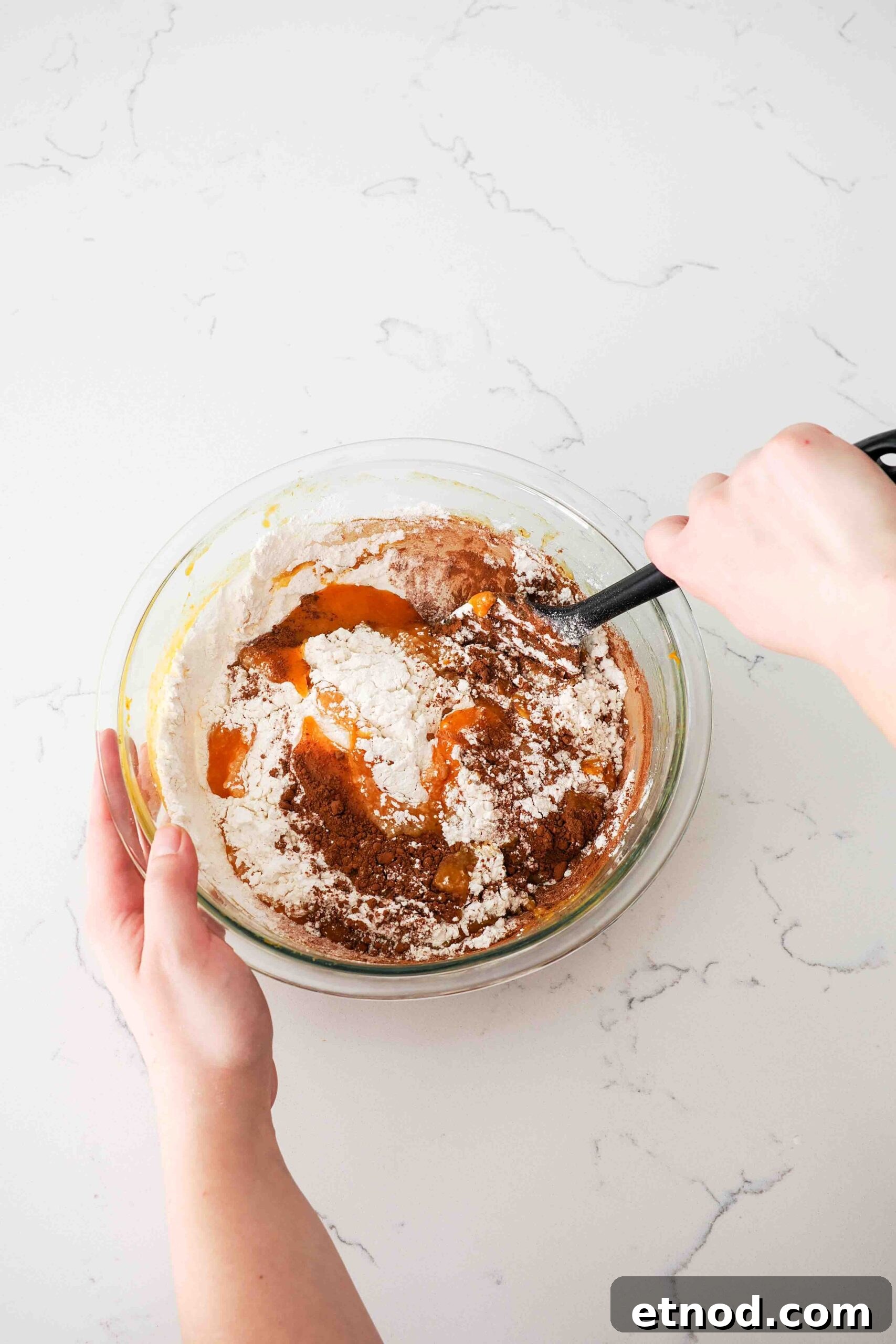 Chocolate Pumpkin Muffins with Melty Chips — Fall Baking Recipe 6 Two hands use a spatula to stir in the dry ingredients into a wet pumpkin batter.