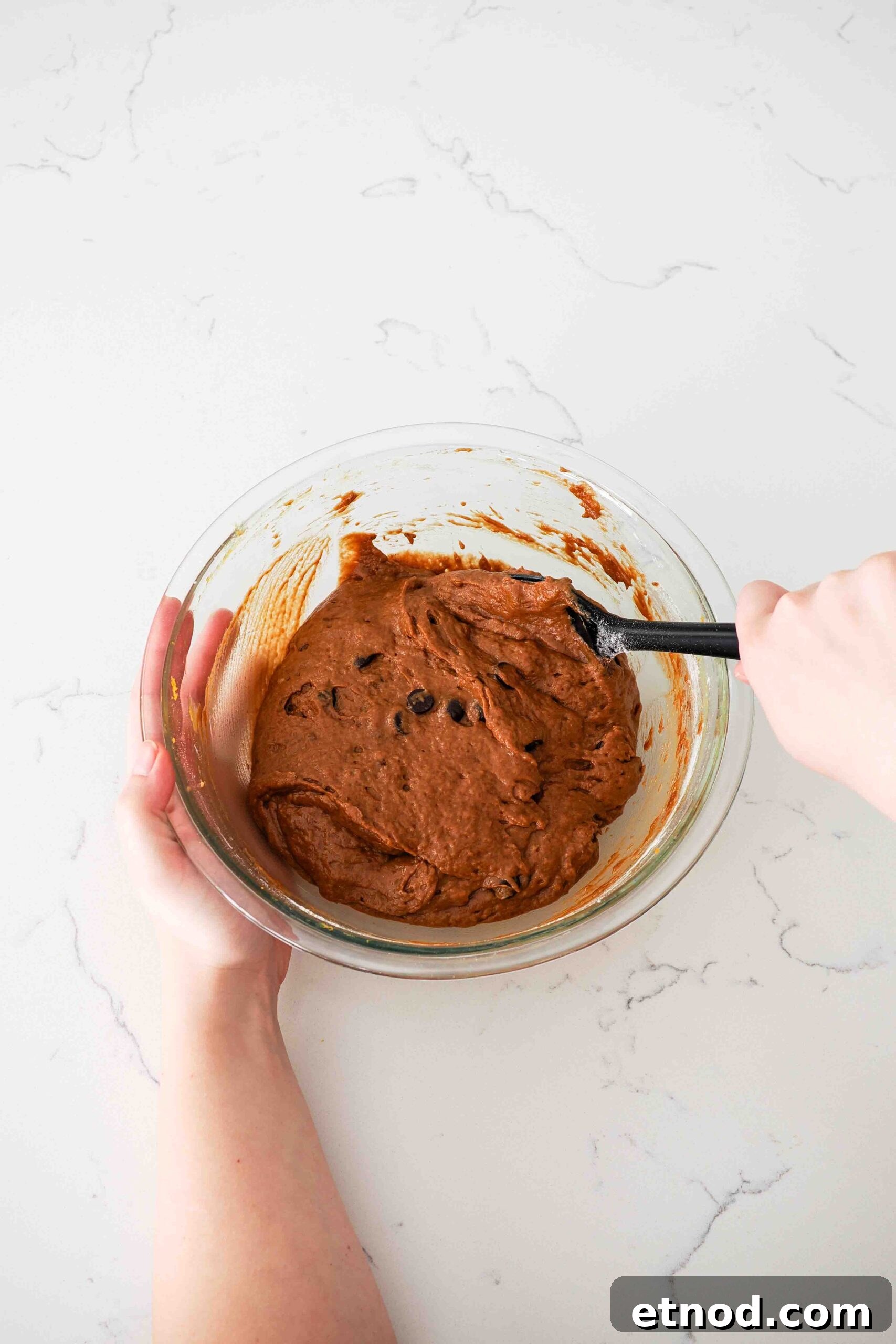 Chocolate Pumpkin Muffins with Melty Chips — Fall Baking Recipe 7 Two hands use a spatula to stir together chocolate pumpkin muffin batter.