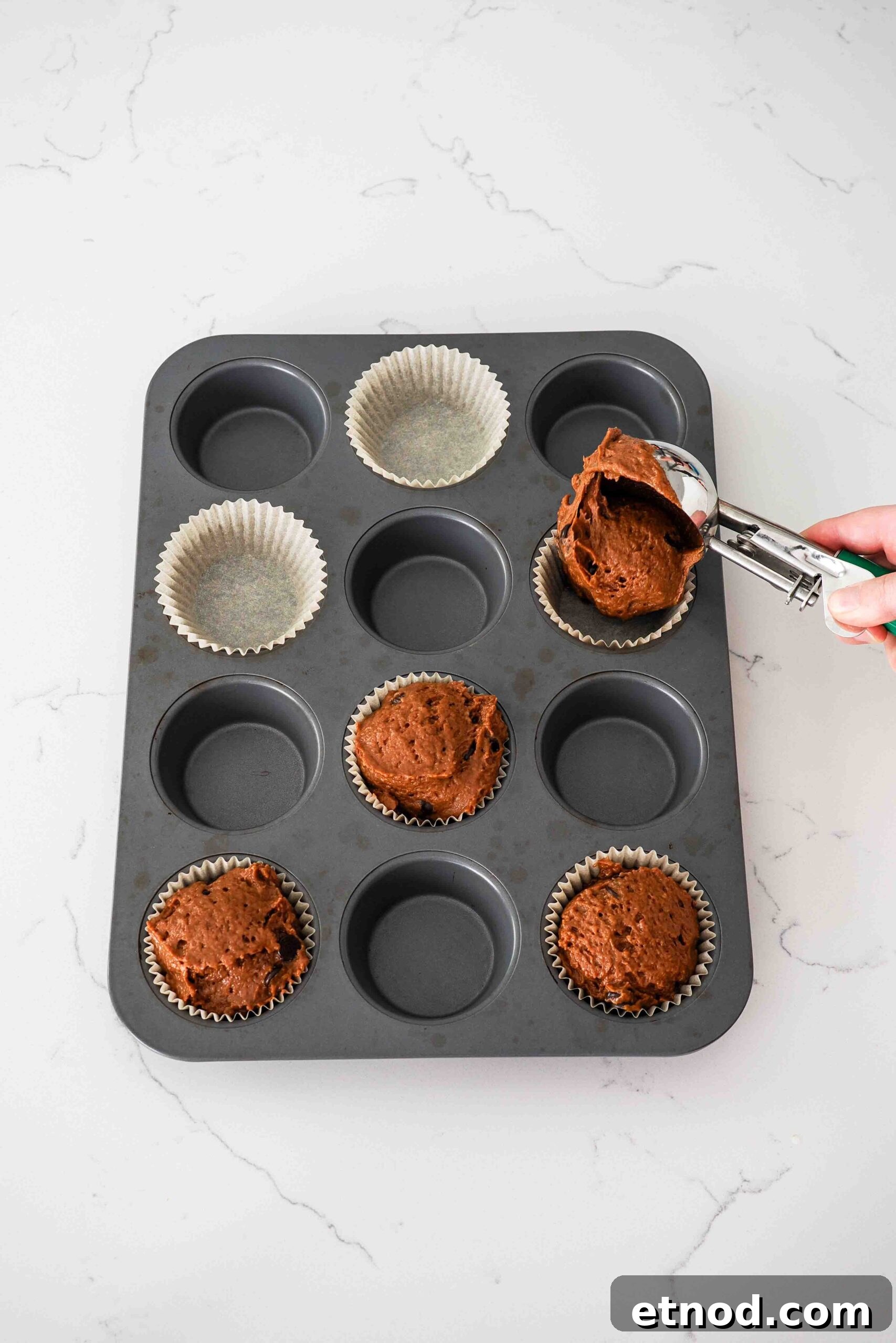 Chocolate Pumpkin Muffins with Melty Chips — Fall Baking Recipe 8 A hand scoops chocolate pumpkin muffin batter into a lined muffin pan.