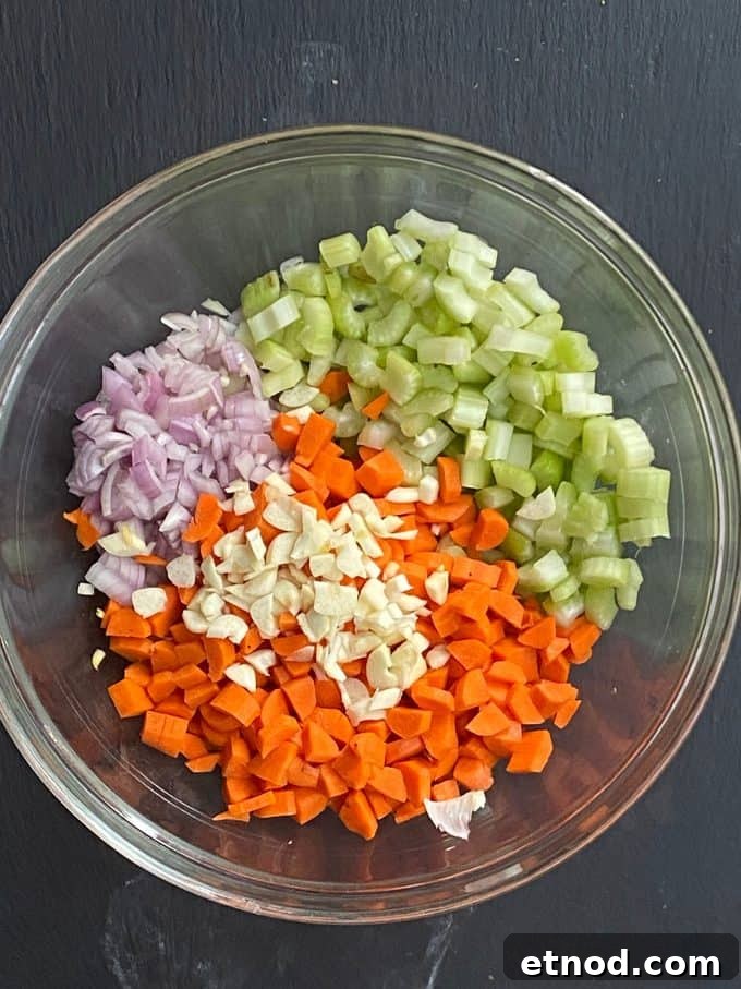 An overhead view of a glass mixing bowl with chopped onion, celery, carrots and minced garlic