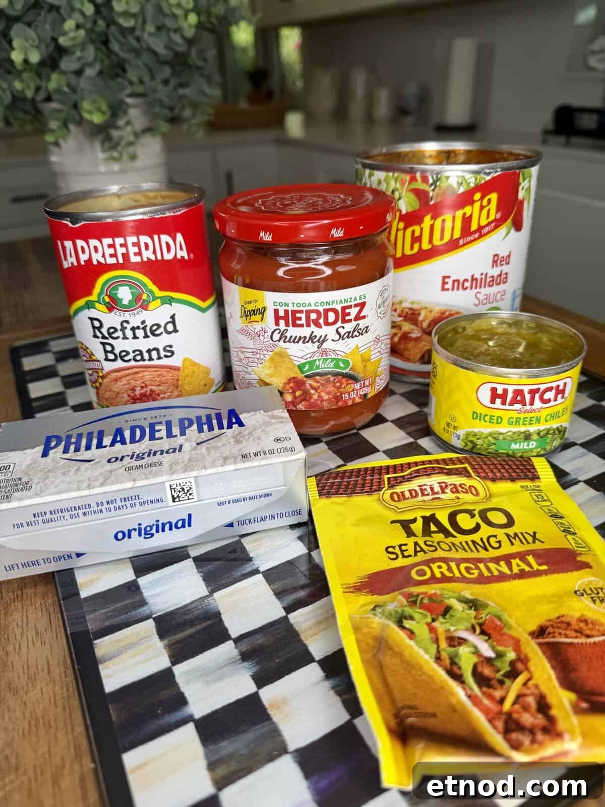 Creamy Baked Salsa Chicken Recipe for Weeknight Dinners 3 A variety of Mexican food ingredients on a checkered surface, including refried beans, chunky salsa, enchilada sauce, diced green chiles, cream cheese, and taco seasoning mix, set in a kitchen.