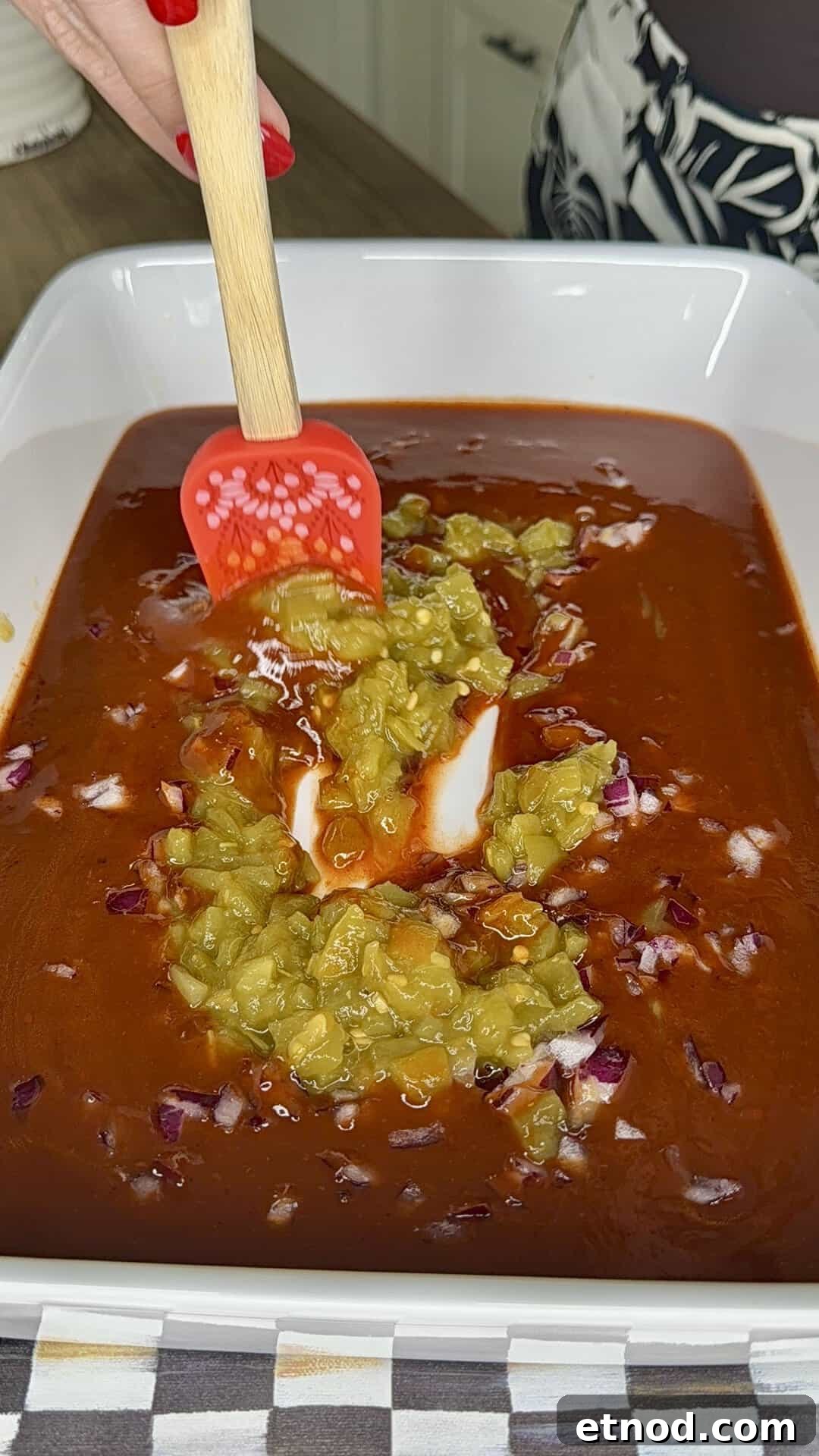 Creamy Baked Salsa Chicken Recipe for Weeknight Dinners 4 A hand with red-painted nails uses a red spatula to mix green chiles, chopped red onions, and red sauce in a white rectangular dish on a kitchen counter.