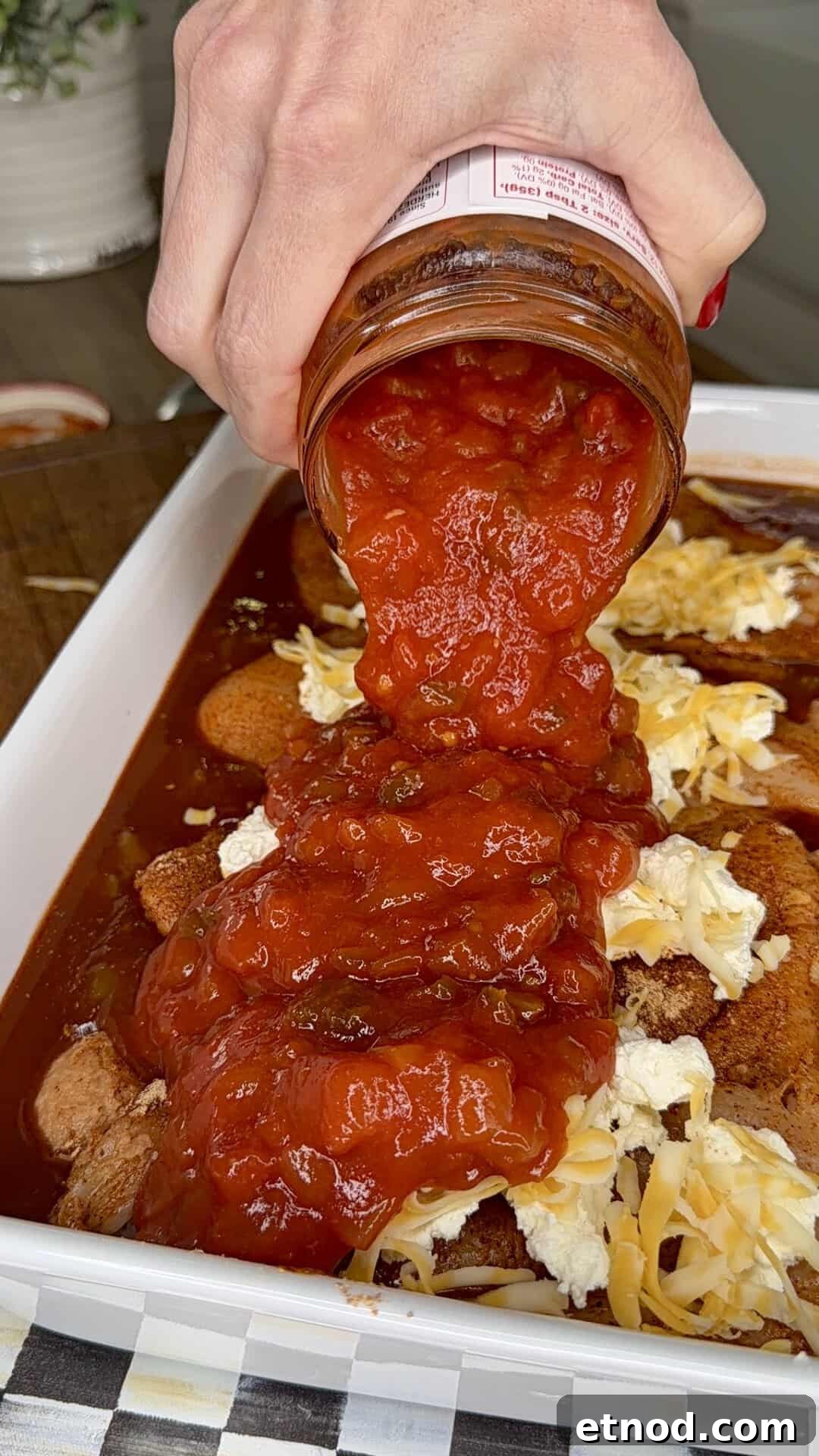 Creamy Baked Salsa Chicken Recipe for Weeknight Dinners 6 A hand pours chunky tomato sauce from a jar over a dish filled with cheese and other ingredients, preparing a meal in a white rectangular baking dish.