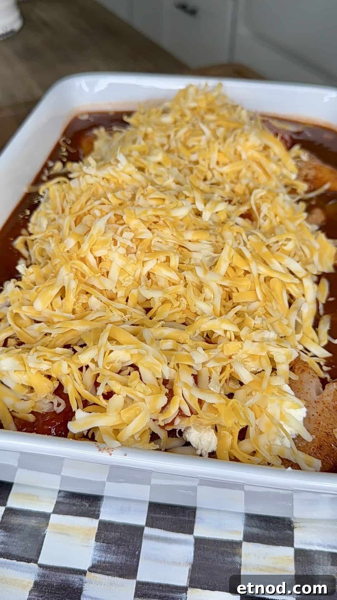 Creamy Baked Salsa Chicken Recipe for Weeknight Dinners 7 A baking dish filled with enchiladas topped with generous amounts of shredded cheddar and Monterey Jack cheese, ready to be baked. The dish sits on a checkered-patterned surface.