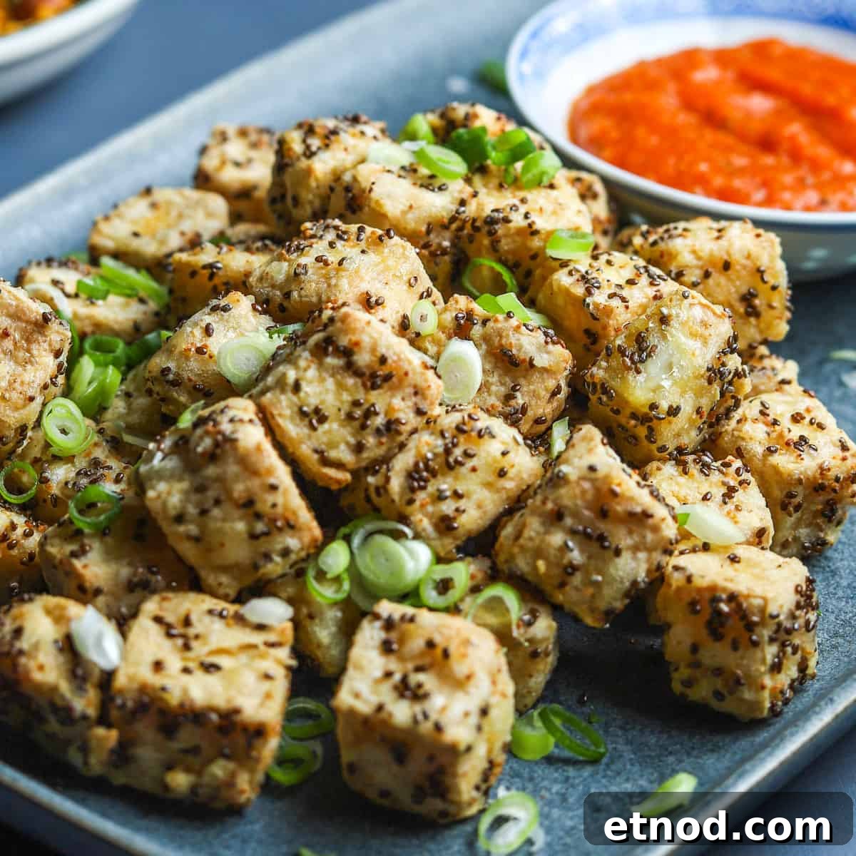 Crispy Air Fryer Tofu Coated with Chia Seeds for Extra Crunch 2 photo of a blue oblong plate filled with crispy air fried tofu with spring onion sprinkled on top and a bowl of kimchi sauce in the background.
