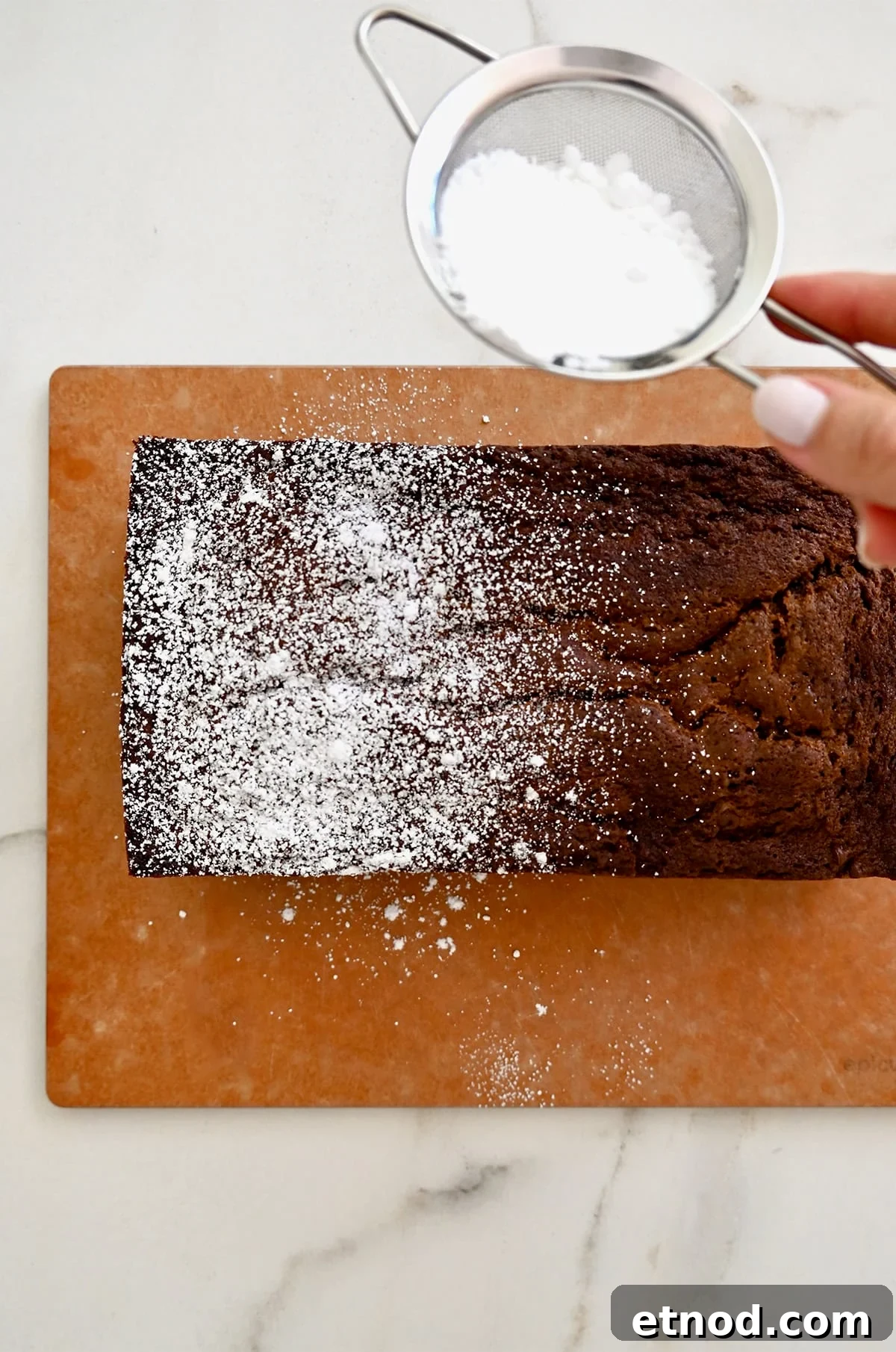 Decadent Chocolate Loaf Cake Recipe for Moist, Rich Results 11 A hand holding a small metal sieve, dusting powdered sugar over the top of a chocolate loaf cake studded with mini chocolate chips.